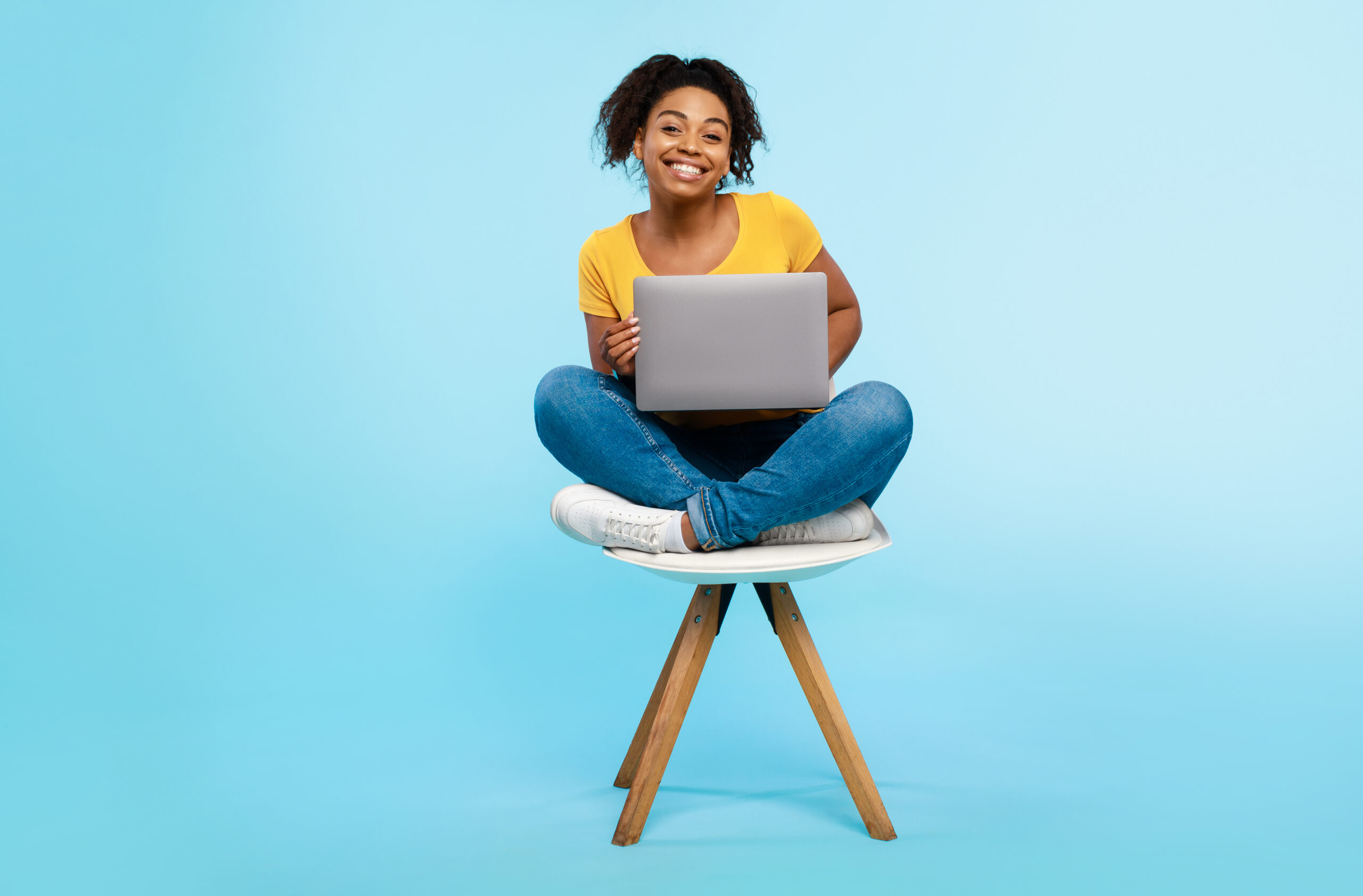 Cheery young black woman working online, sitting on chair and using laptop on blue studio background, full length Two friends smiling